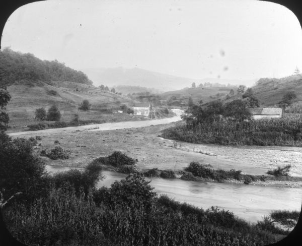 View of a rural landscape with a river running through it. Farm buildings on either side.