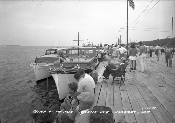 View of a crowd gathered along a pier for Regatta Day. Boats are docked alongside the pier on the left.