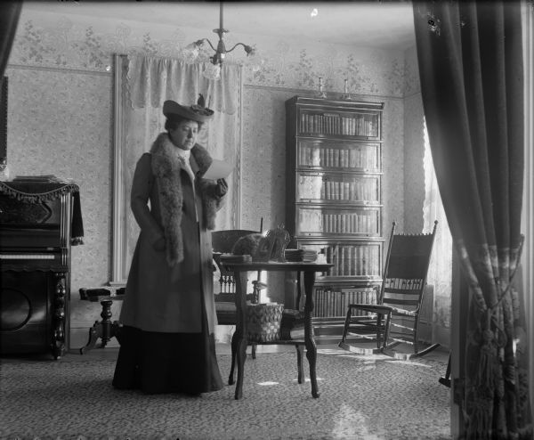 Mrs. A.G. Zimmerman posing in a home on W. Washington Avenue. She is standing reading a document in a parlor with a piano.