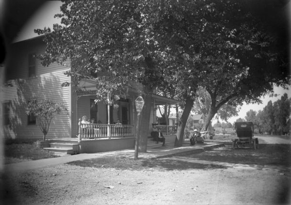 Exterior view of the Riverside Hotel, North 1st Street opposite the railroad depot. Two women, one holding a dog in her lap, and a man are sitting in chairs on the porch. A couple is sitting on the front steps, and there is a man sitting in a hammock set up next large wooden walkway in front of the hotel which has trees growing through it. An automobile is parked in the street.