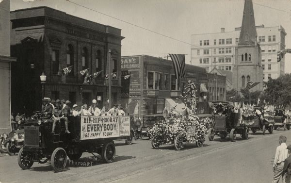 A Fourth of July parade on S. Carrol Street featuring decorated vehicles. Flags are hanging from the sides of commercial buildings.