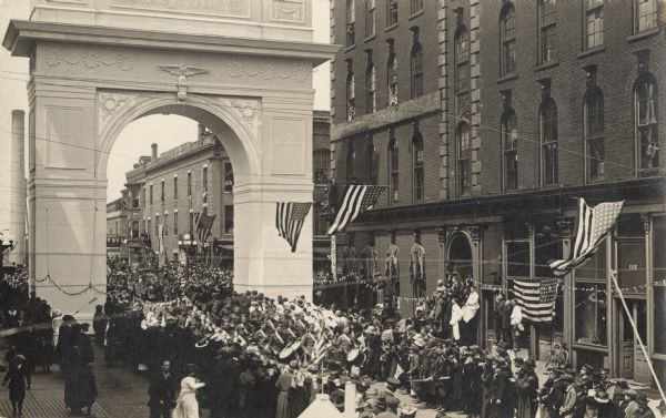 Elevated view of a crowd lining the street near the "Welcome Home" Arch anticipating a parade of returning veterans. The facades of the buildings are decorated with flags.