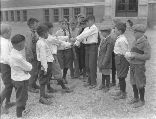 Outdoor view of a group of boys about to play softball, and using a bat to choose who goes first. Location identified as Emerson School. A dog is standing in the background near the school building.
