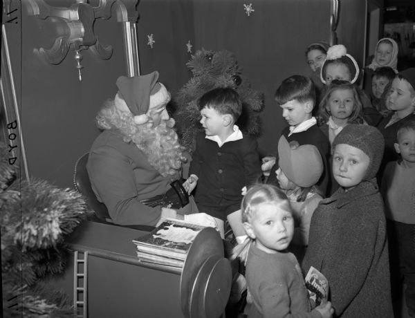A group of children meeting Santa at Gimbels. Decorated Christmas trees are next to his chair.