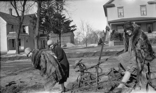 Two women are bending over in front of the plow on the left, pretending to pull a woman sitting on the back of the plow which is parked near a residential street. All three women are wearing hats and coats, two of fur, and dresses. There are houses in the background.