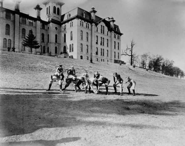 The Whitewater State College football team posing in a snap formation on a hill just outside the college. People are watching from a college building at the top of the hill in the background.