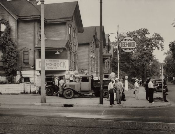 View across street towards Harry Rodin's service station at the intersection of North 7th and West Galena. People are standing at the corner near the bus stop.