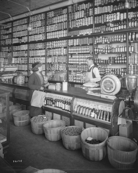 A woman is being waited on by a man standing behind the counter of a grocery store. Canned goods are stacked on shelves along the wall behind the counter.