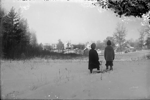 Winter scene with Muriel and Laurie Peterson, the photographer's children, reflecting on the snow conditions near their home. A group of houses are in the distance. A sled is in the snow behind them.