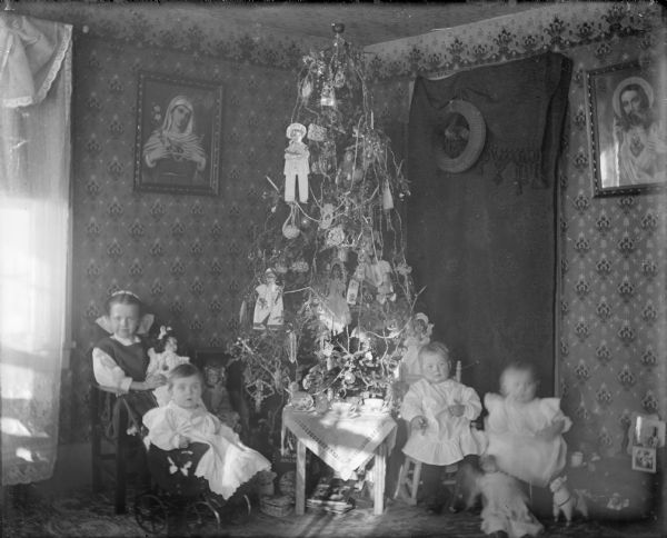 Group portrait of the Ponkratz girls sitting around the Christmas tree. Two girls are sitting on the left, with the older girl holding a doll, and the younger girl sitting in a baby carriage. The other two girls are sitting on the right, with a doll on the floor at their feet. In the center in front of the tree is a table set with china.