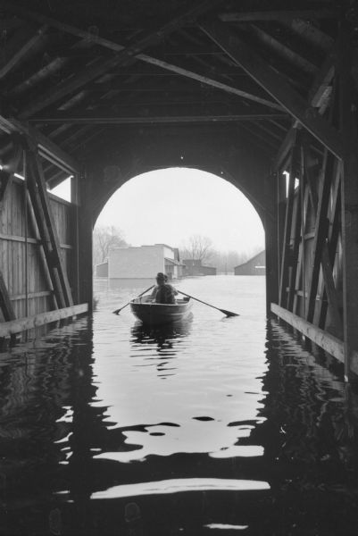 A man and another person are in a rowboat in the flooded waters under the entrance to the covered bridge in Stonefield Village, which is a Wisconsin Historical Society site.