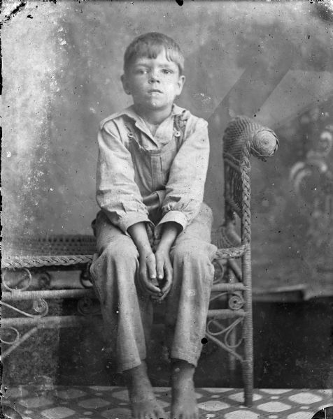Studio portrait of Earl Lester sitting on a wicker bench in front of a painted backdrop. He is wearing a shirt and bib overalls.