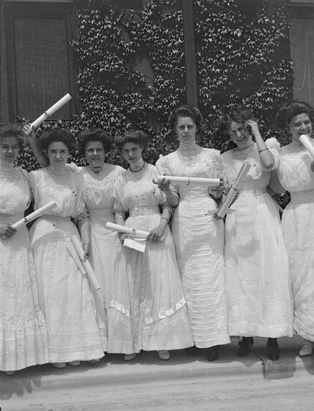 Outdoor group portrait of women graduating from Downer College standing in front of a vine covered wall. The woman are wearing long white dresses with high lace collars, and are each holding a diploma. Nieta Oviatt Friend is third from the left.
