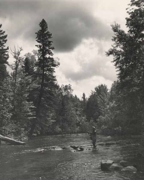 View across water towards a man fly fishing while standing in the river. He is wearing waders and a hat, and a creel and a net are hanging at his sides. He has a pipe in his mouth.