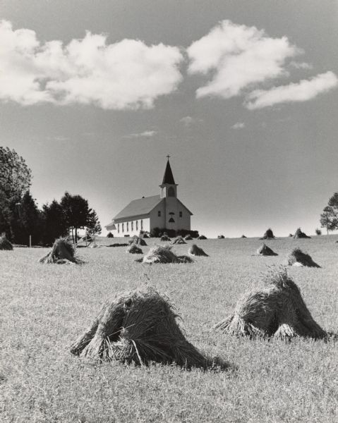 A church with a dark steeple is set on top of a hill above a field with shocks of grain. Trees are on the left and right.