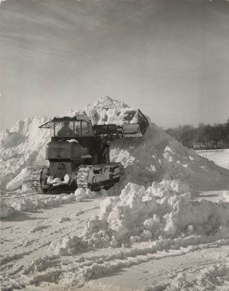 A man is operating an Allis-Chalmers HD 5 Diesel tracked front end loader that is scooping snow onto a large pile. Trees are in the background.