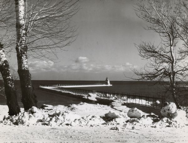 The Manitowoc Breakwater Lighthouse entered service in 1918, was automated in 1971 and sold in 2009. The lighthouse is located on the end of a 400 foot breakwater. Snow and trees are along the Lake Michigan shoreline in the foreground.