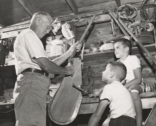 Two young boys observing as an older man is tightening a bolt on the tiller of a sailboat. Tools and other objects are hanging on the wall behind them.