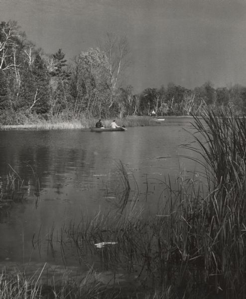 View from shoreline towards two men in a canoe near Birch Point on Little Elkhart Lake. The day is overcast. Around the point is another man in a boat, and tipis on the far shore in the background. The lake is lined with trees and marshy areas.
