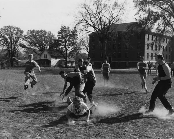 Students playing a game of touch football on a dirt field at Norbert College. One team is wearing shirts with "Inmates" printed on the front, other shirts have "Warriors."