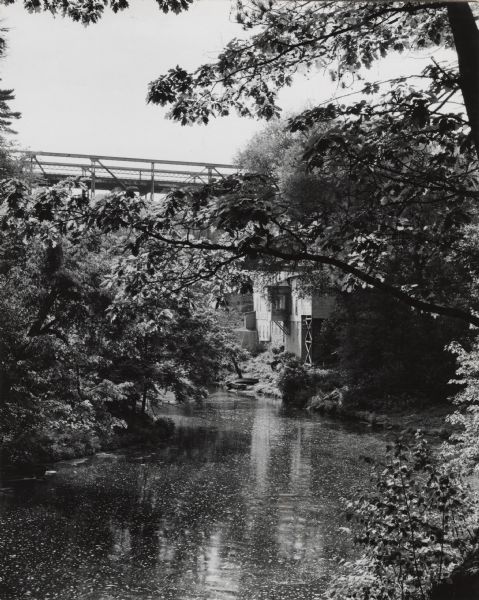 Mirror Lake, below the dam at Timme Mill. Trees and foliage cover the banks and overhang the water. The one lane high reverse metal truss bridge, commonly called "the Upside Down Bridge," was built in 1908. The bridge was replaced by a two lane concrete bridge in 1986.