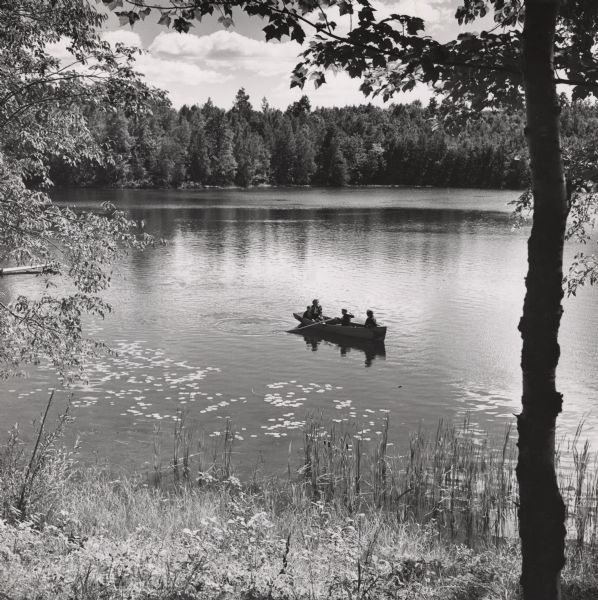 Tree-lined view from shoreline looking down at a lake scene of two adults and two children in a rowboat. Lily pads are floating on the surface of the lake near the shoreline. There is a pier on the far left, and in the background is a tree-lined shoreline.