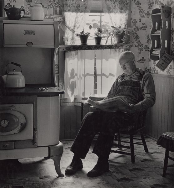 Lawrence Sherman sitting in a chair by the window, reading the paper in his farm kitchen. On the left is a Jungers Model E stove with kettles and a teapot. Behind him is a Royster's Almanac hanging from a bookshelf on a string. Plants are lined up on a shelf in the window.