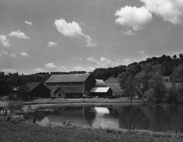 Rural scene of a cluster of farm buildings with a pond in the foreground. On the horizon, trees cover the hills.
