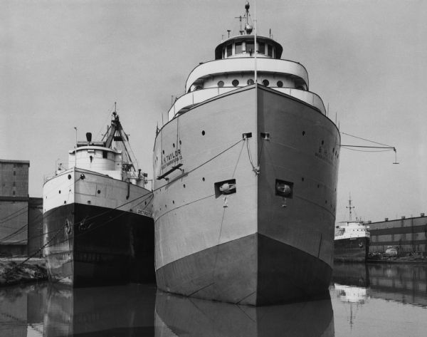 Three lake steamers docked in the harbor. The closest steamer is the B.H. Taylor of the Bradley Transportation Company, transporting ores. Buildings are in the background.