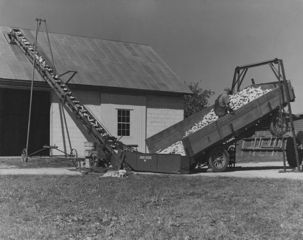 Robert Palzer, owner of the farm, is using a Model 5-C John Deere portable farm elevator to convey hybrid corn to the kernel remover in the building. The words "John Deere," "Bridge Trussed" and "Non Rust" are on the elevator. The wagon is tilted using a wagon lift. Mr. Palzer is in the wagon box. A farm dog is resting in the grass.