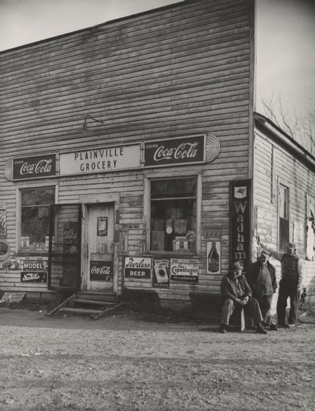 Three men visit at the corner of the Plainville Grocery. There are many signs and logos on the storefront: "Coca-Cola," "Peerless Beer," "Model Smoking Tobacco," "Copenhagen Chew," "Wadhams Sinclair," "Bire-ley's Orange," "Dad's Root Beer," "Chesterfield," "Lucky Strike," "Nesbit's Orange," "7-Up," "Old Dutch Cleanser," "Leverich for State Senator," and "Philip Morris."<p>Comment from photographer: "This old country store is typical of so many of the Adams County stores. I only wish it were possible to photograph the inside."</p>