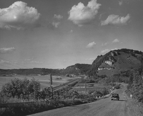 View of an automobile traveling north on Highway 35, near the Mississippi River. Railroad tracks run between the road and the river. In the background are tree covered bluffs. A sign on the right reads: "Side Road."