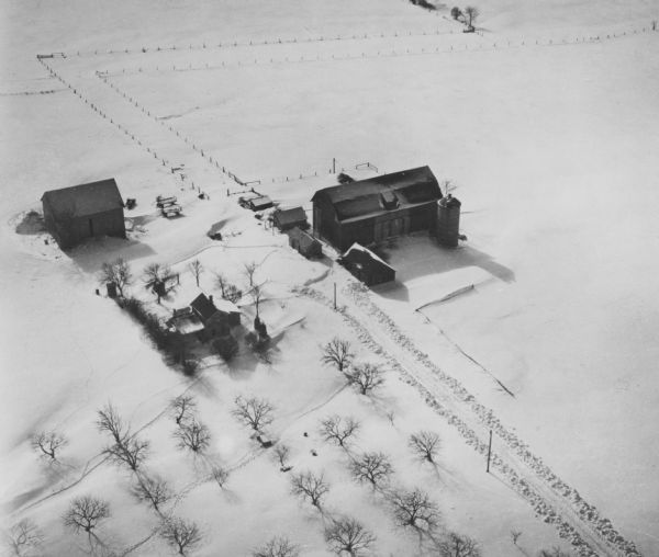 Aerial view of a farm in mid-winter. Snow blankets the ground and some of the buildings. An orchard is in the foreground.