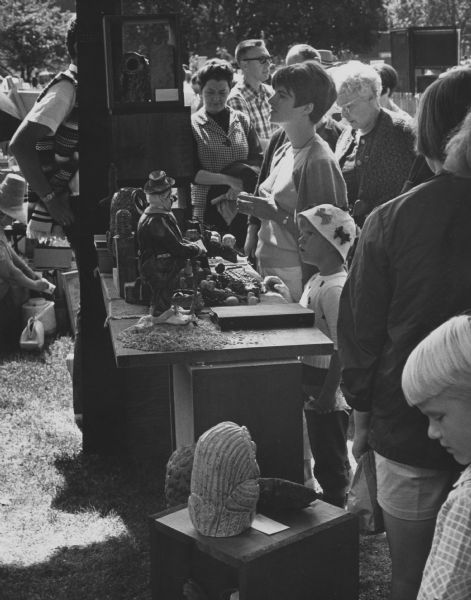 A crowd browsing the art at a fair in a small town. The booth is displaying mostly owls and may belong to artist Clarence P. Cameron.