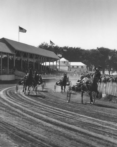 Harness racing at a County Fair. The horses pulling sulkies are making the turn in front of the grandstand. Another building and trees are in the background. Spectators are also watching from the infield on the right. Four American flags are flying over the buildings.