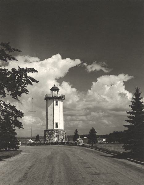 View down road towards the Fond du Lac lighthouse, built in 1933. There are cumulus clouds in the sky, and pine trees are lining the shore.