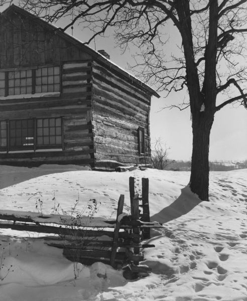 Unidentified log house in winter, located near Highway 83. A fence is in the foreground and a tree on the right.