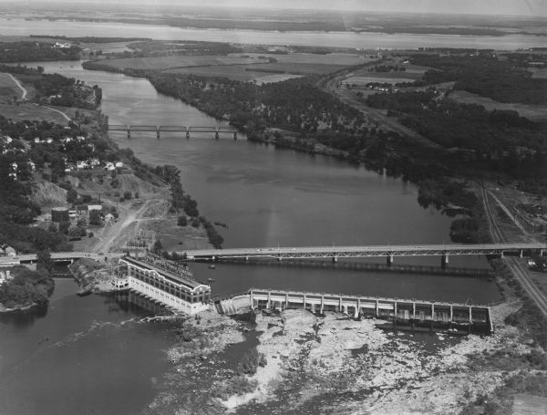 Aerial view of a hydroelectric power plant owned by Northern States Power Company located on the Chippewa River. A highway bridge and a railroad bridge span the river.