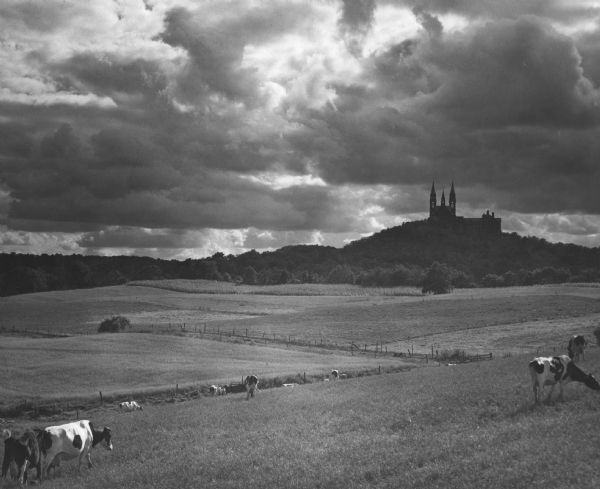 Dramatic view of Holy Hill against a sky full of clouds. In the foreground, cows are grazing in fenced fields.