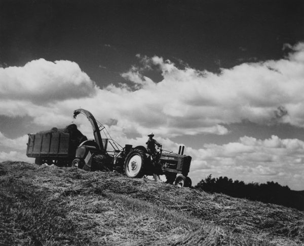 Haying on the farm with dramatic clouds in the background. The John Deere tractor is pulling the chopper as it empties into a wagon. 