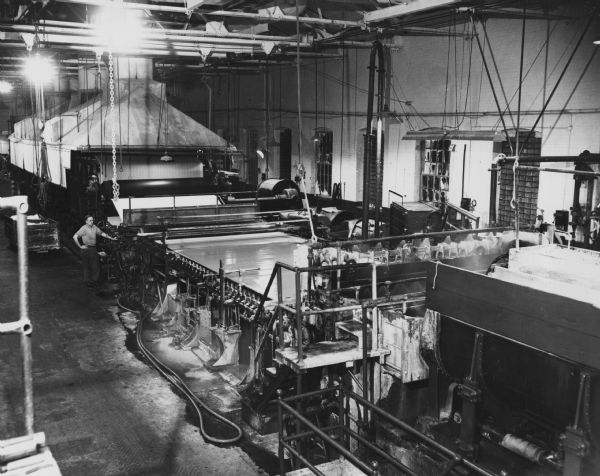 Elevated interior view of a paper mill with paper making machinery. An employee is standing next to the machinery on the left.