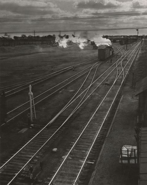 Elevated view of parallel tracks in a railroad yard, with a locomotive pulling a boxcar. A man is standing near the boxcar. On the right are buildings, and on the left is a switch. In the distance are more trains waiting on the tracks.