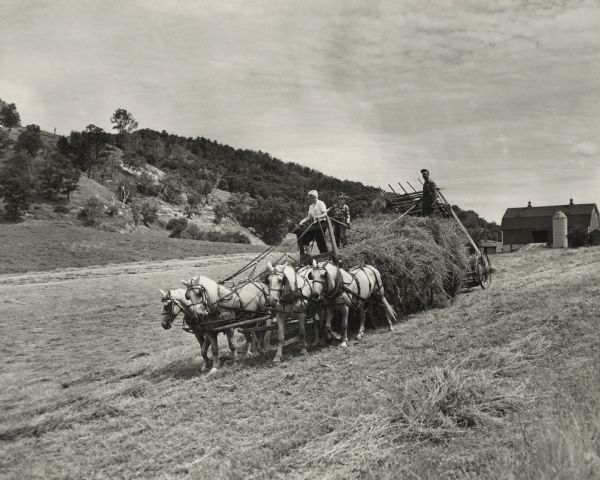 A woman is driving a horse-drawn hay wagon pulling a hay loader on the William Houser Farm. A child and a man, each with a hay fork, are riding on the load. On the right is the barn, on the left are the bluffs that enclose Sand Lake Coulee.
