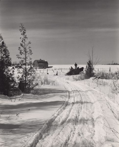 Two men hauling logs in a sled on a snow-covered farm. Farm buildings are in the distance, with trees on the left and right in the foreground.