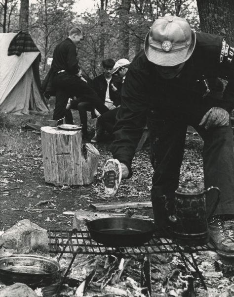A Boy Scout or Scout Leader, with "Madison Wis 101" on his shoulder patch, making pancakes in a skillet over an open fire. Beside the skillet is a coffee pot. A plate, fork and small pan are resting on a stump. Scouts and a tent are in the background. Caption on the reverse reads: "Camp Cooking. Klondike-Sauk County."