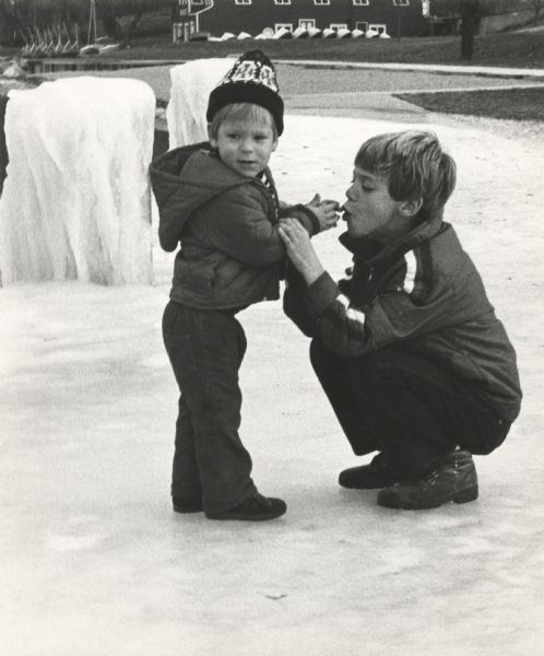 A boy is blowing warm air on his little brother's hands to warm them up. They are standing on ice at James Madison Park. In the background are boats pulled up on the shore for the winter.