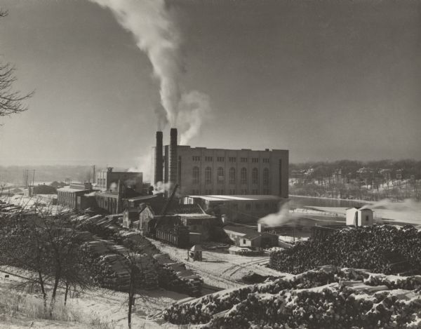 Winter image of the Interlake Pulp Mill in the Fox River Valley. The buildings are surrounded by large stacks of logs. The Fox River can be seen behind and to the right of the main building.