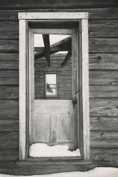View of trees and a farm building as seen through a dilapidated door and window of an old house. Snow is on the threshold of the door and in front of the building. All the wood is weathered and the roof is missing.