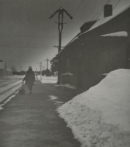 A passenger waiting for a train in front of the Chicago and Northwestern railroad station on a clear and cold evening. The person is holding a lamp, and a satchel is on the ground at their feet. Overhead is a semaphore above the station, and in the right foreground is a snowbank.