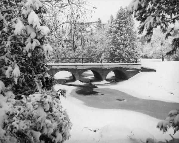 Snow scene of a concrete bridge with three arches, over a creek, in Silver Creek Park. The creek is mostly frozen and surrounded by snow covered trees.